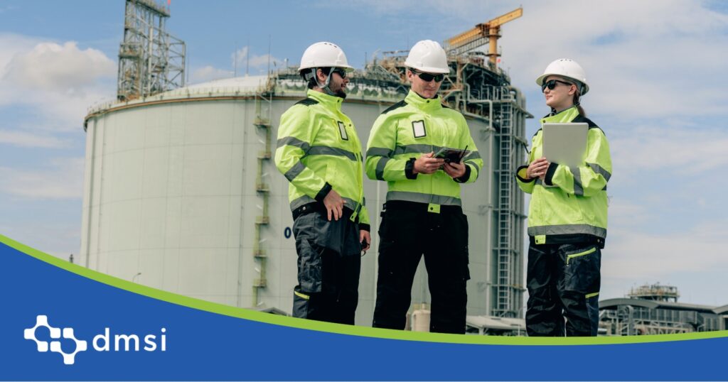 Three field technicians in hi-vis jackets using tablets to review equipment data outside a large industrial storage facility