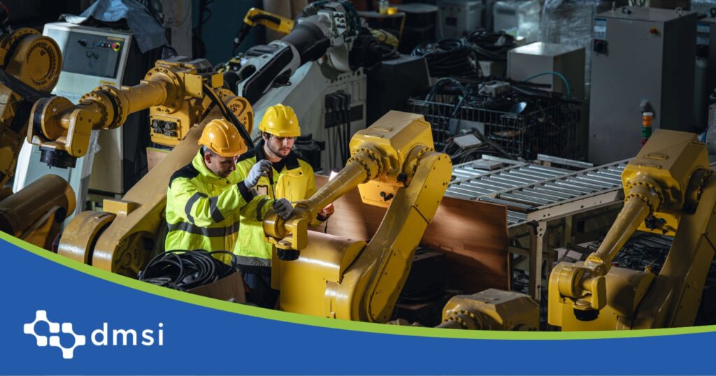 Two maintenance workers in hard hats and hi-vis jackets inspect a yellow industrial robotic arm.
